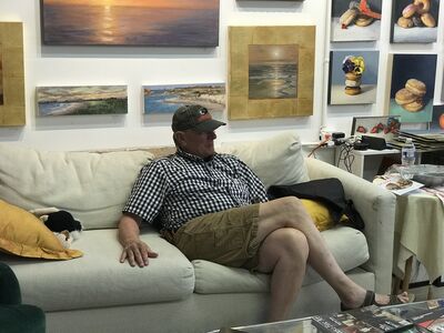 Keywords: artist,seated,couch,older man,cap,black and white shirt,khacki shorts,artwork,paintings