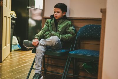 Keywords: boy,seated on a chair,green jacket,tennis shoes,grey trousers,door,wood floor