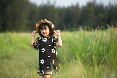 Keywords: girl,green field,beige hat,black dress with white flower,trees in background,arms up