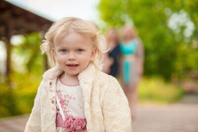 Keywords: child,girl,kid,blonde with short hair,white furry coat,white dress with pink roses,people in the background